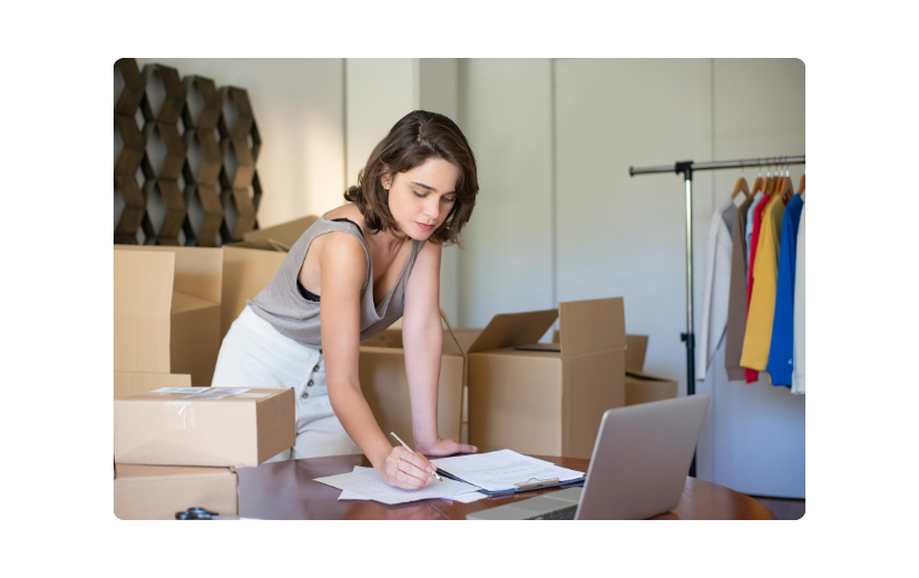 Woman managing an eCommerce business, reviewing documents with shipping boxes and a laptop, focusing on content creation.